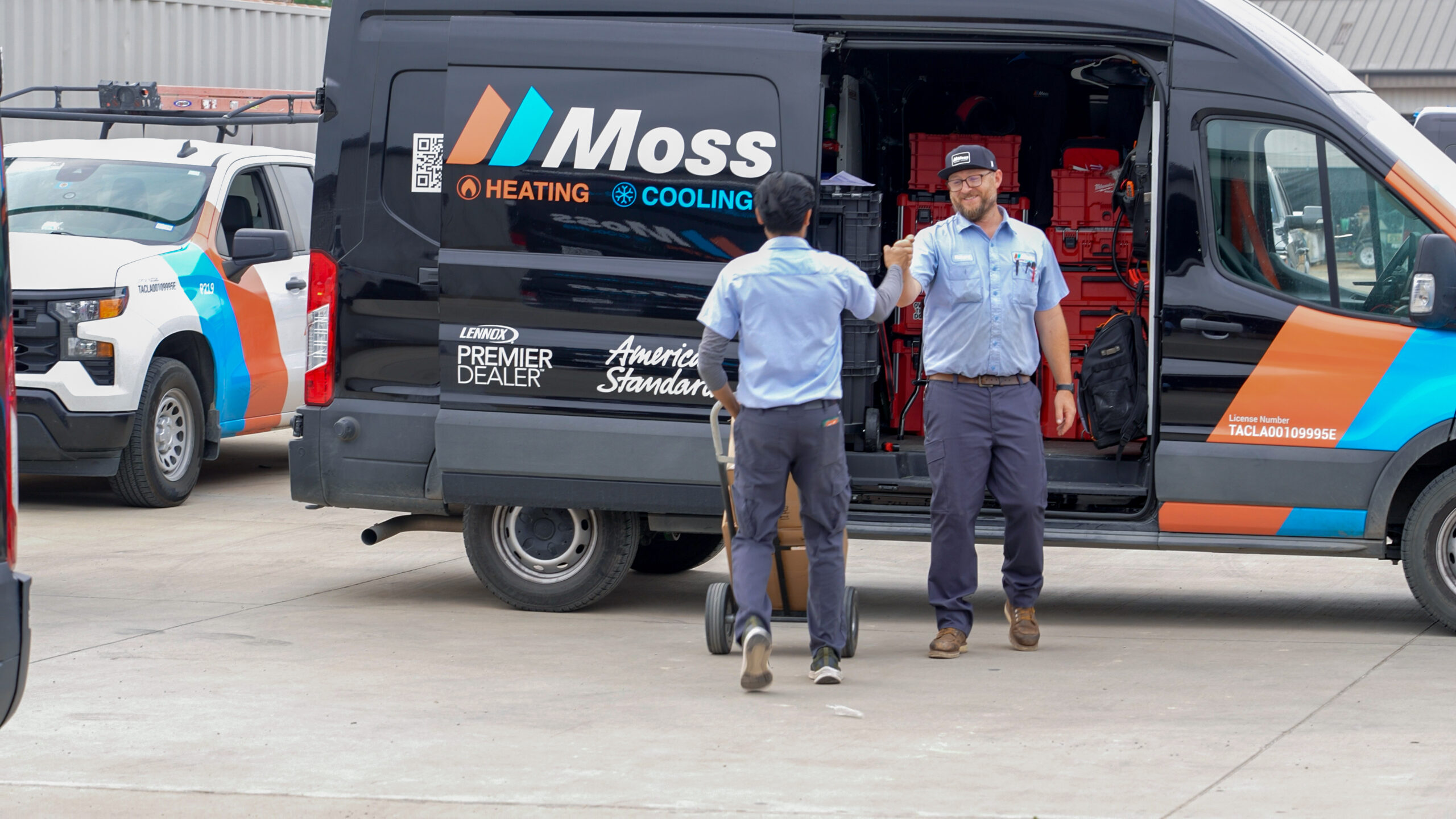 Two technicians in blue uniforms fist-bump beside a Moss Heating and Cooling van with the side door open.