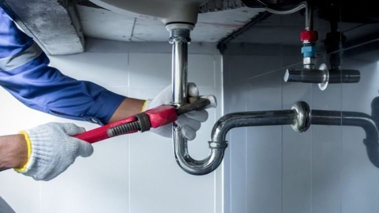 Plumber in gloves uses a red-handled adjustable wrench to tighten a U-shaped pipe under a sink.