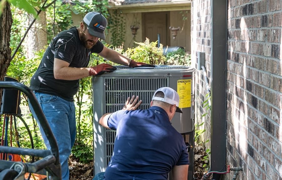 Two HVAC technicians service an outdoor condenser unit beside a brick house.