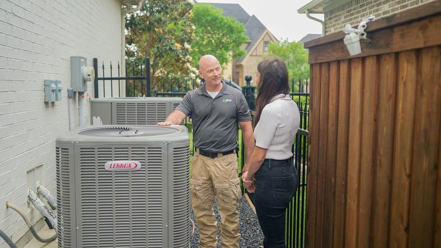 HVAC technician in a gray polo talks with a woman beside an outdoor Lennox air conditioner unit in a fenced backyard.
