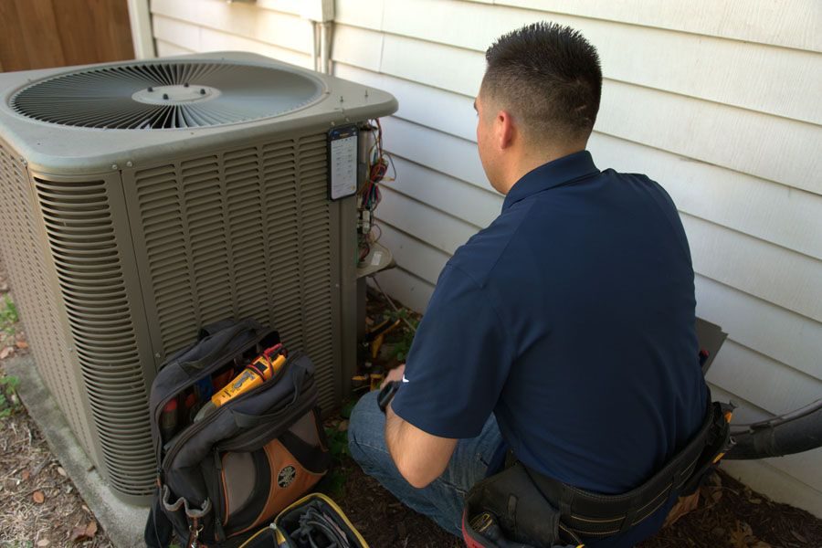 HVAC technician crouches beside an outdoor air conditioner unit, working with tools from a bag on the ground.