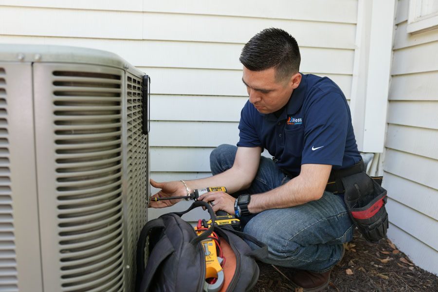 HVAC technician kneels beside an outdoor air conditioner unit, using tools on a condenser panel.