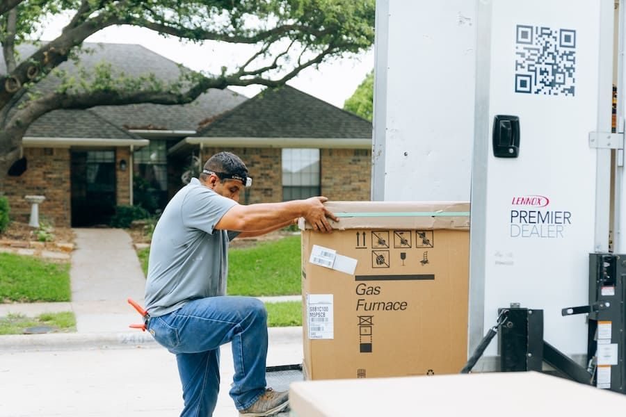 Man in a gray shirt lifts a large box labeled 'Gas Furnace' from a moving truck in a suburban street.