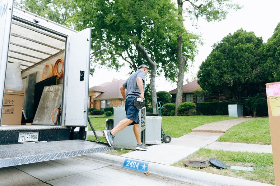 Man in shorts unloads boxes from a moving truck into a suburban driveway.