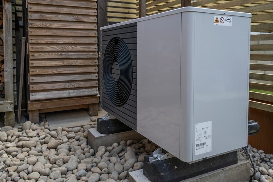 Outdoor heat pump/air conditioner condenser unit mounted on concrete blocks beside a wooden fence, resting on gravel pebbles.