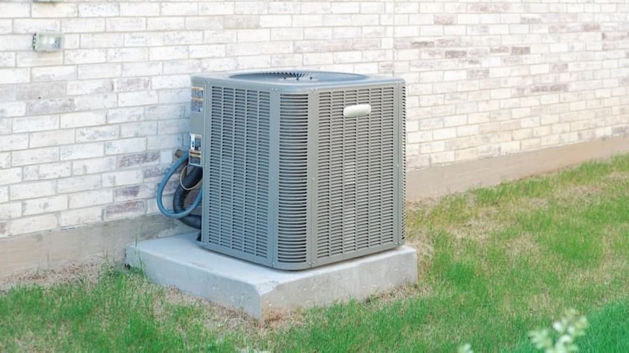 Outdoor air conditioning condenser unit on a concrete pad beside a brick wall, with green grass nearby.