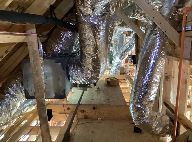 Insulated flexible ductwork runs along wooden attic joists with an exposed air handler and unfinished ceiling panels nearby.