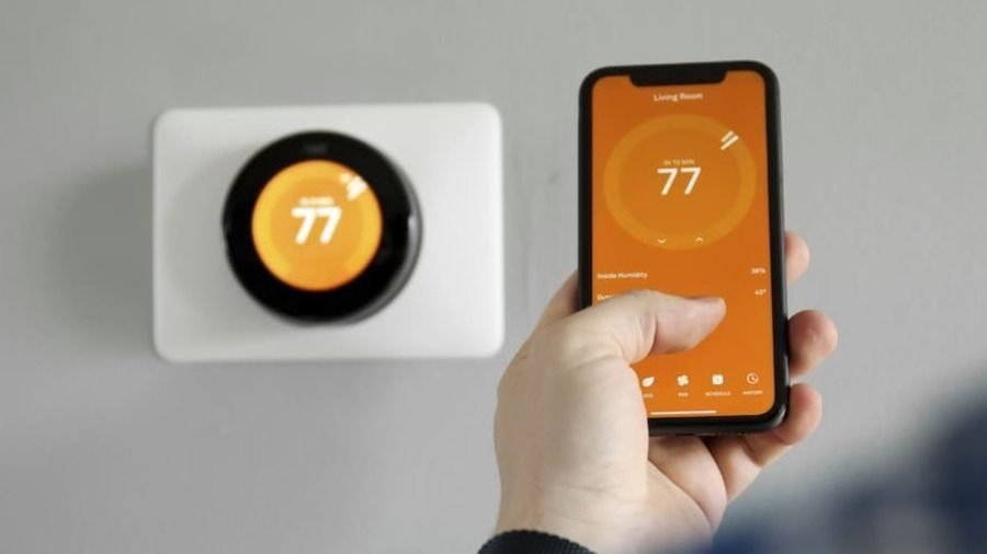 Hand holds a smartphone displaying a smart thermostat app at 77°F; wall thermostat shows 77.