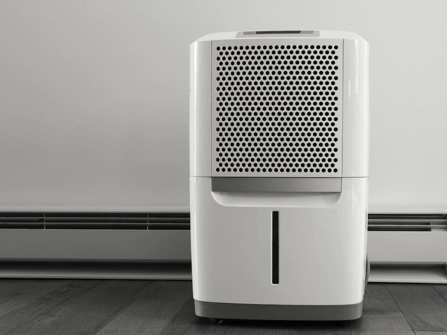 White portable dehumidifier with a perforated front panel sits on a dark floor against a light wall, featuring a water level window at the bottom.