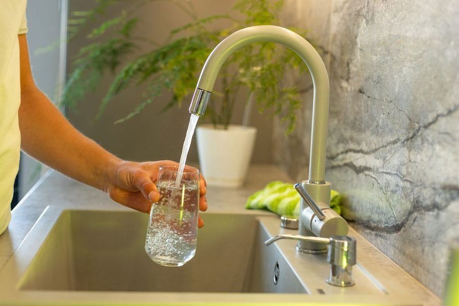 Person rinsing a glass under a modern kitchen faucet with running water above a sink.