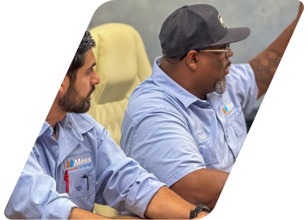 Two coworkers in light-blue work shirts with Moss logos sit side by side, looking toward a speaker in a meeting room.