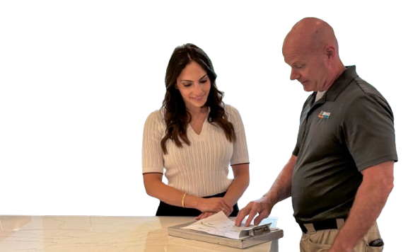 Woman in a white knit top signs documents on a clipboard at a reception counter as a man in a polo observes nearby.