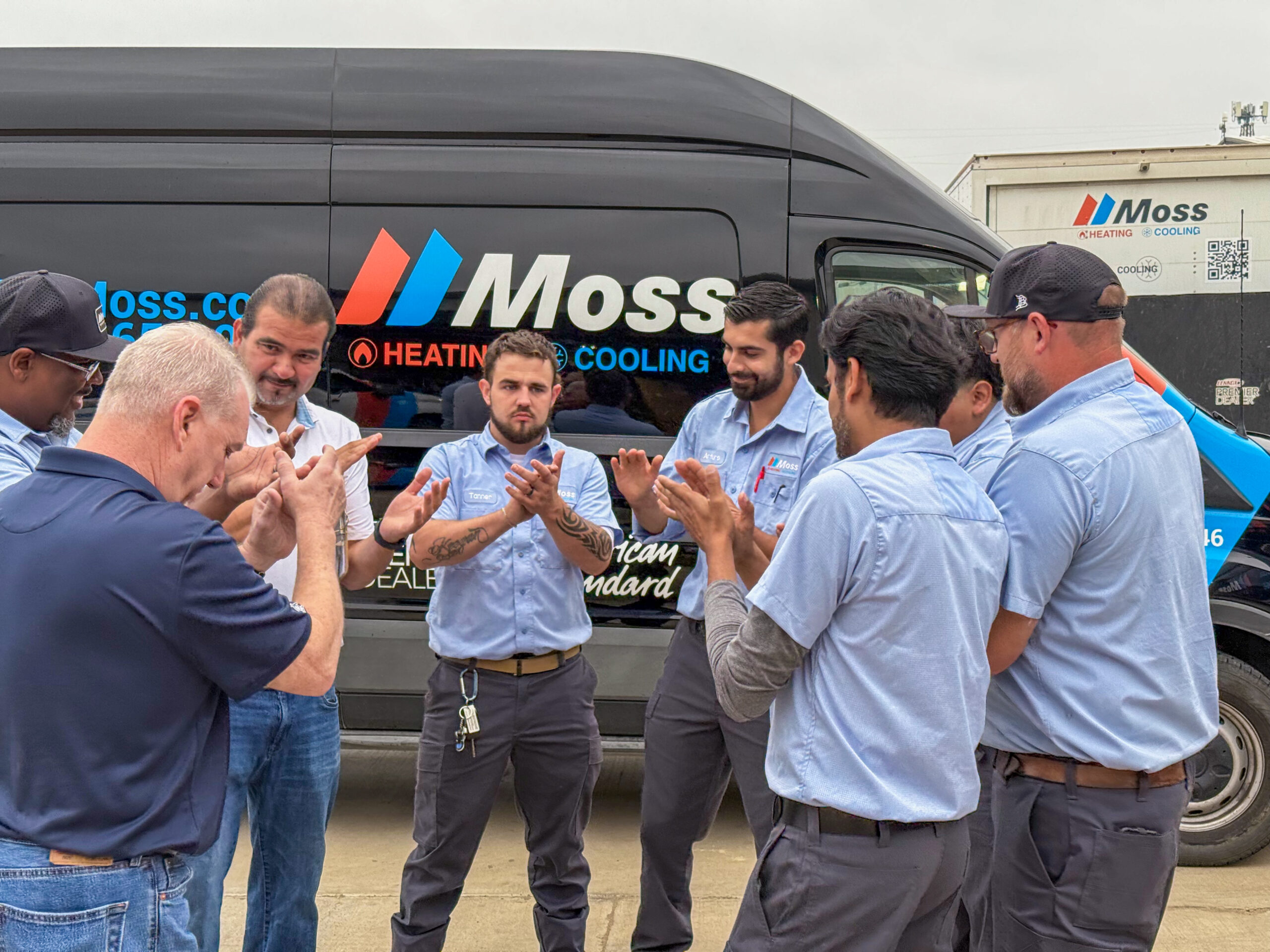 Group of service technicians in blue shirts stand in front of a Moss van, clapping and smiling together.