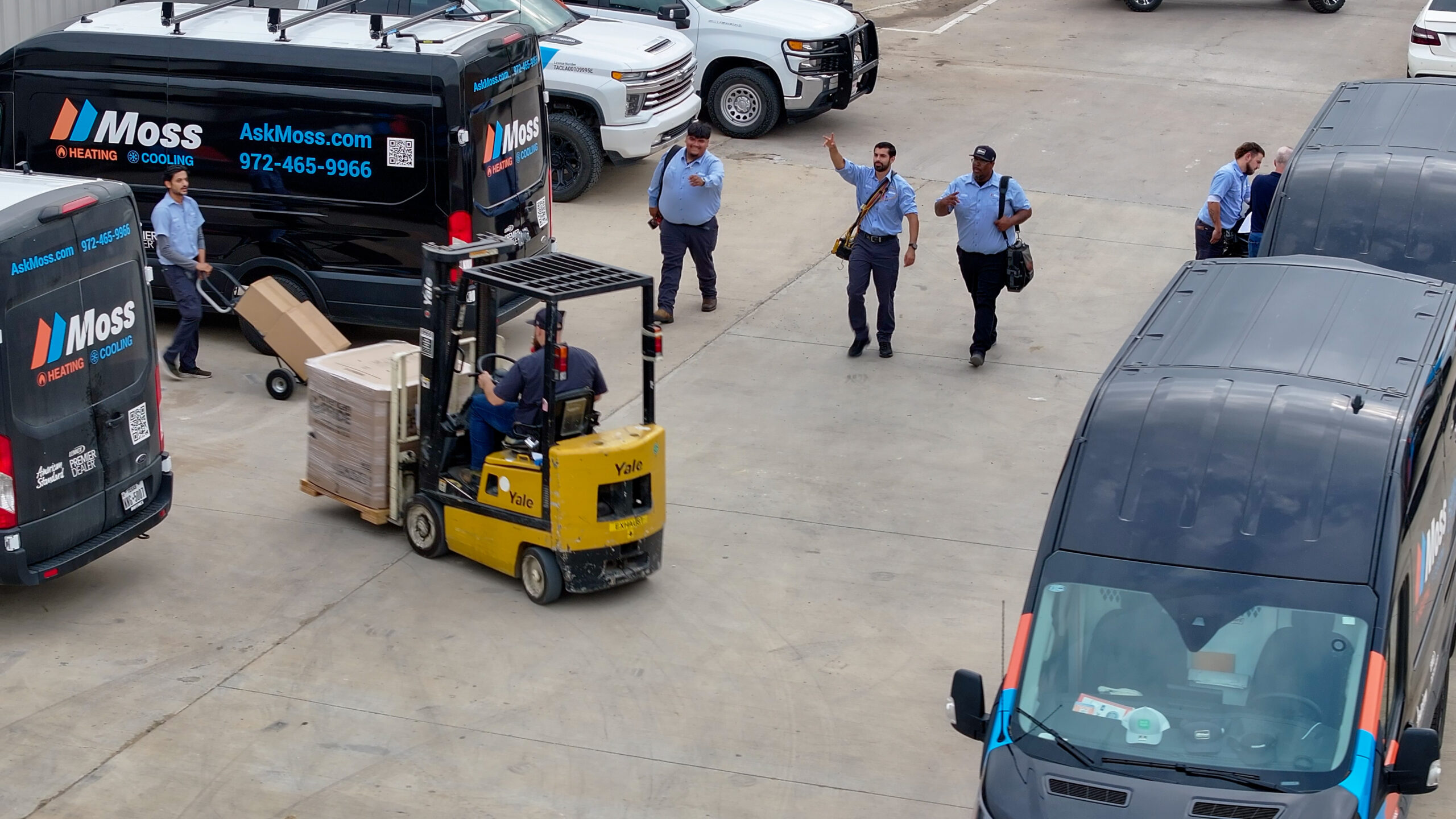 Parking lot scene with Moss Heating & Cooling vans and workers guiding a forklift carrying a pallet of boxes toward a truck/fleet area of the yard.