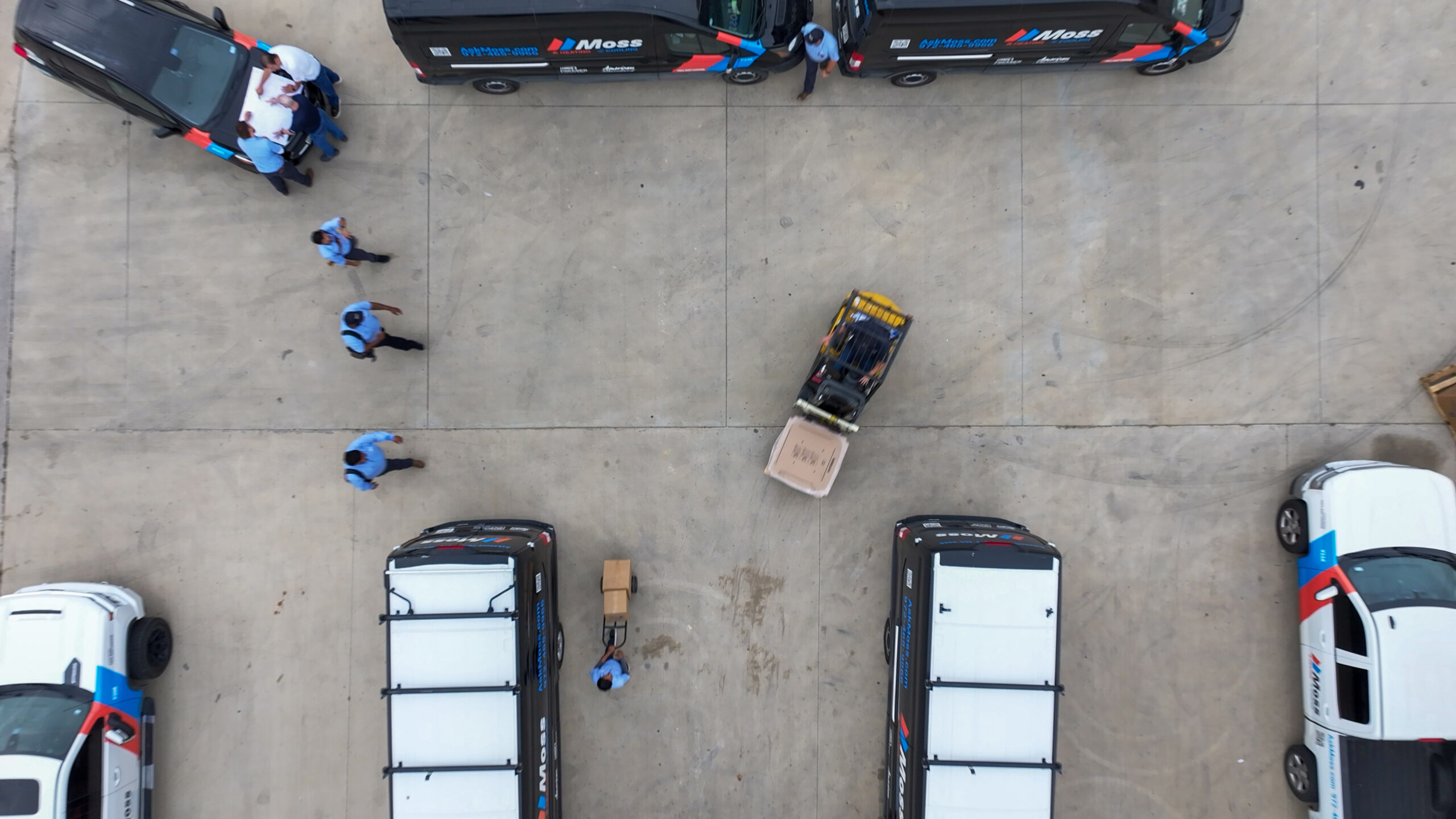 Aerial view of a service yard with Moss-branded vans, workers in blue shirts gathered around a table on a van hood, and a yellow pallet cart nearby.