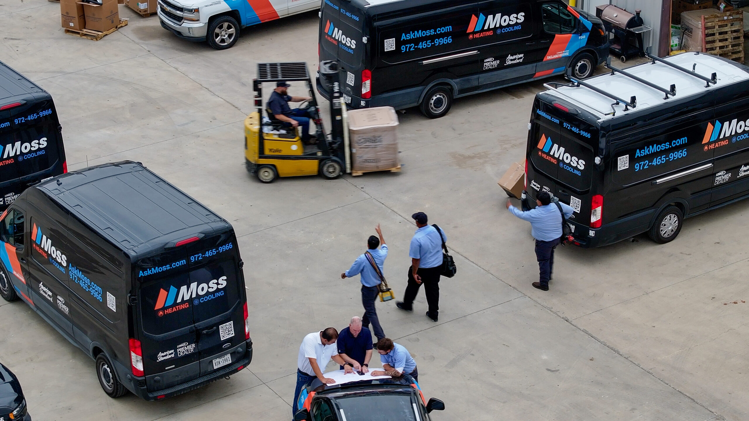 Aerial view of a loading yard with several Moss Heating & Cooling vans, a yellow forklift moving a large pallet, and workers in blue shirts gathered around a table with papers.