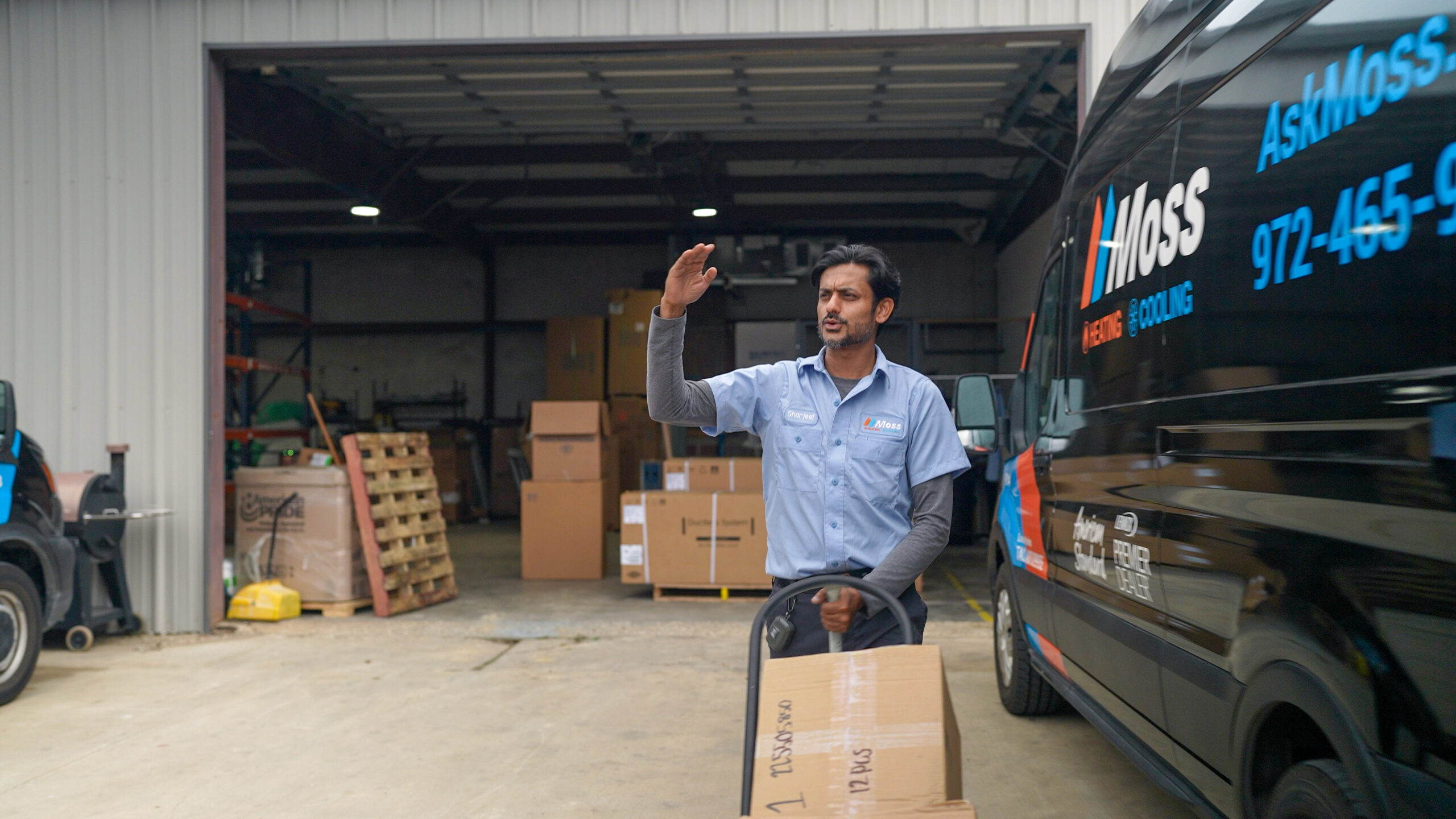 Delivery worker in a light-blue shirt waves beside a Moss-branded van at a warehouse loading area, with boxes and pallets in the background.