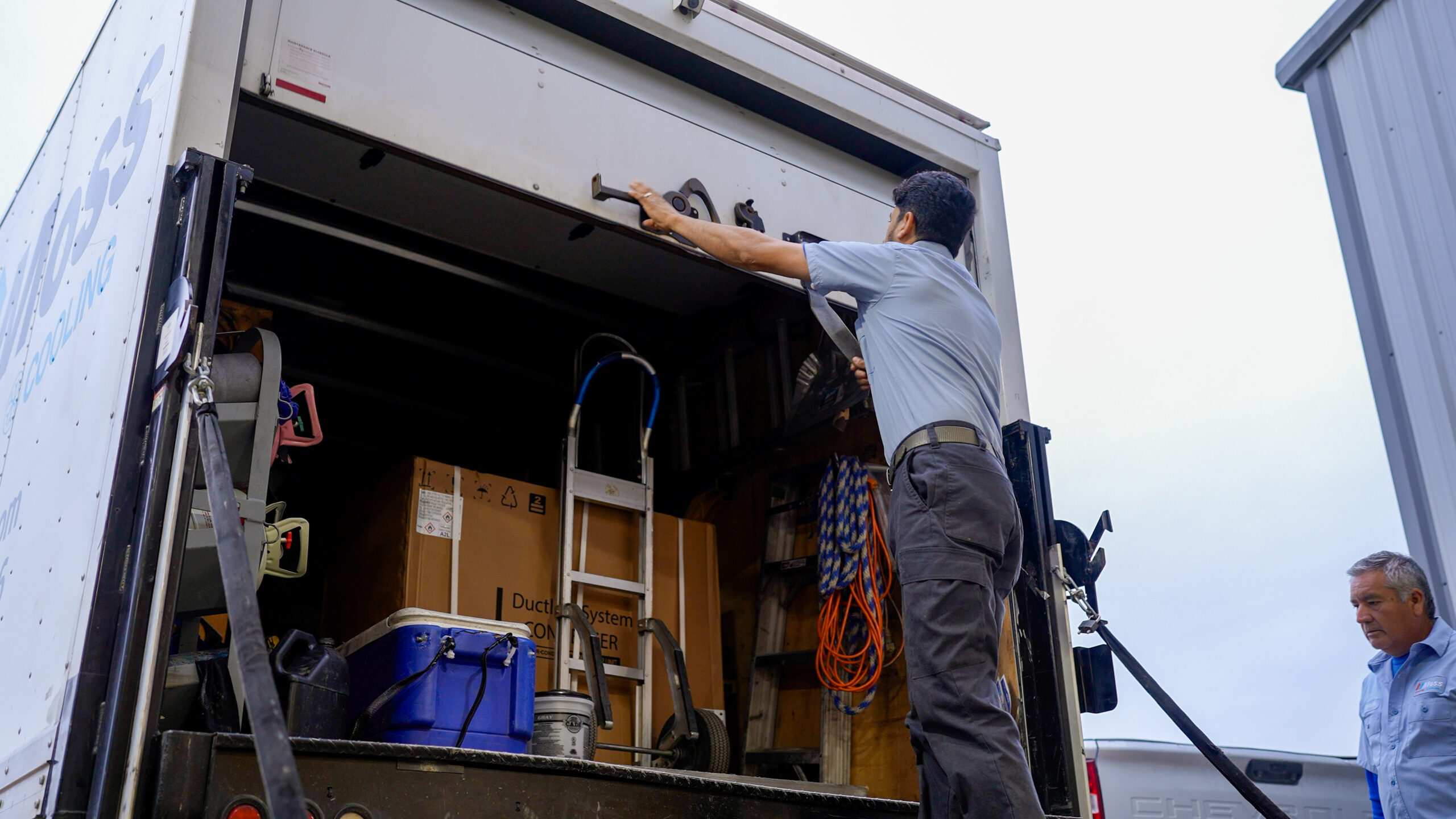 Worker in a blue shirt reaches into an open back of a large delivery truck to secure cargo, with boxes and gear inside.
