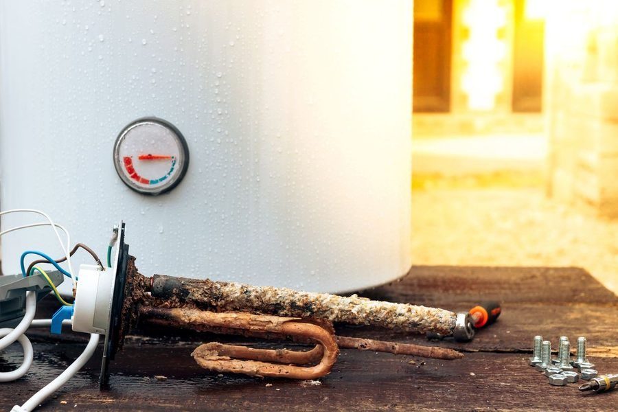 Old, corroded water heater element with exposed rusty pipes and wiring on a wooden surface, outlets and screws nearby.