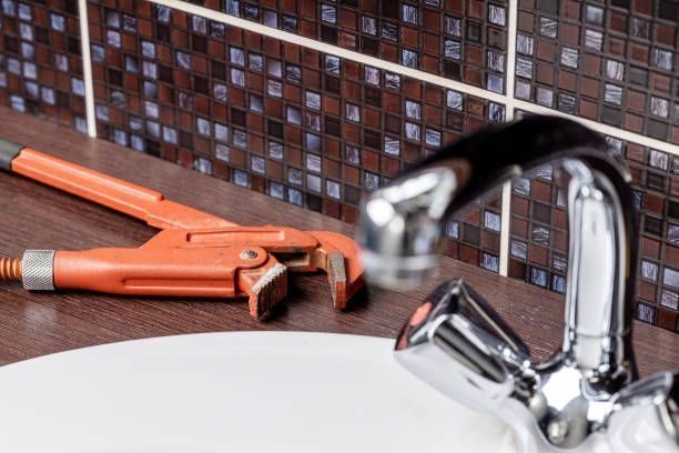 Orange pipe wrench resting on a wooden vanity next to a chrome bathroom faucet above a white sink, with a tiled backsplash in the background.
