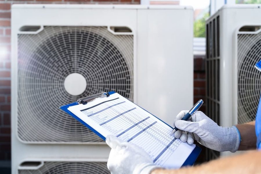 HVAC technician in gloves holds a clipboard and inspects an outdoor air conditioner unit","A technician checks a service sheet on a clipboard beside two large outdoor AC units","Gloved worker writes on a clipboard beside outdoor HVAC equipment during maintenance","Maintenance worker with clipboard evaluates readings near air conditioning units outside a building","Clipped form and pen used by a technician to record data beside street-side AC condensers
