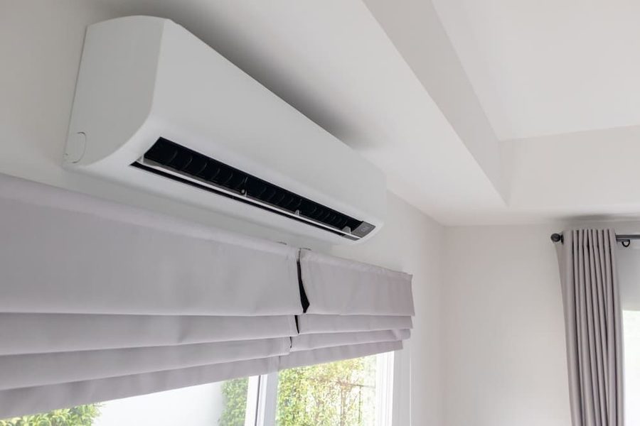 White wall-mounted split air conditioner unit above large window with horizontal blinds partially open, cooling the room.