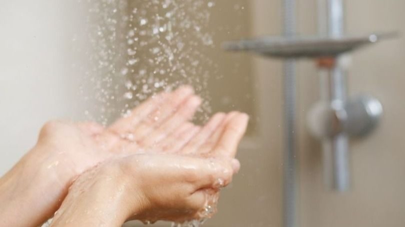 Hands under running water in a bathroom with a shower faucet visible in the background.